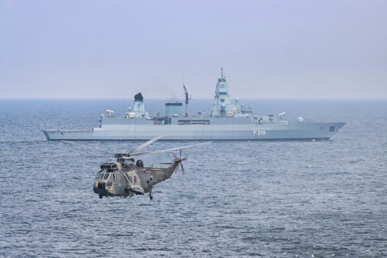 A military helicopter flies over the sea near a German warship, F219.